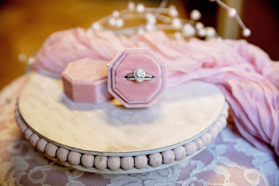 Pink velvet ring box with a diamond ring on a beaded round pedestal with lace and a blush cloth backdrop