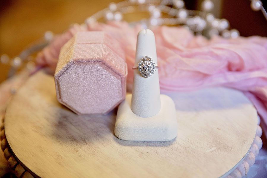Diamond ring ring on a stand beside a pink faceted box and blush ribbon on a tray
