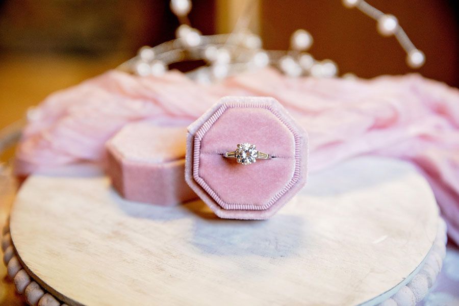 Pink velvet ring box with a diamond ring on a round tray, softly lit against a blurred background