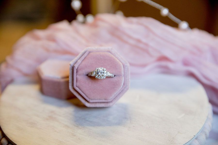 Engagement ring in a pink velvet box on a round marble stand, with soft pink flowers in the background