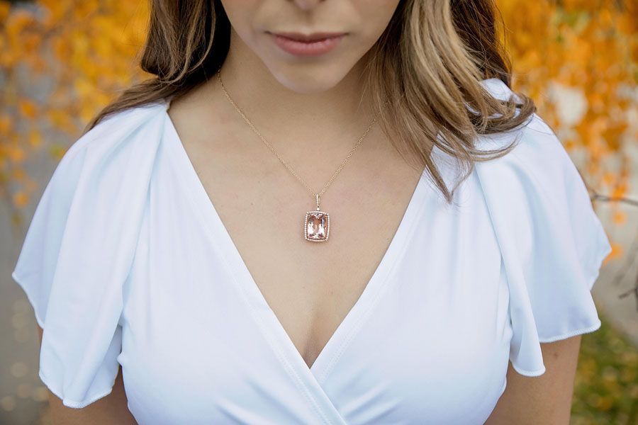 Woman in a white V-neck dress wearing a pink pendant necklace against a blurred autumn background