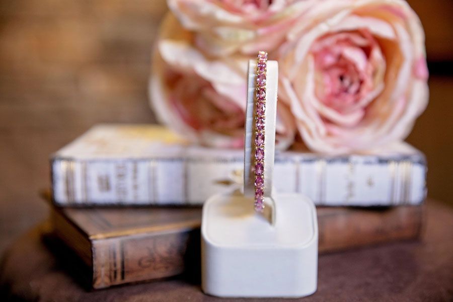 Pink jeweled ring displayed on a white stand with roses and stacked books in the background