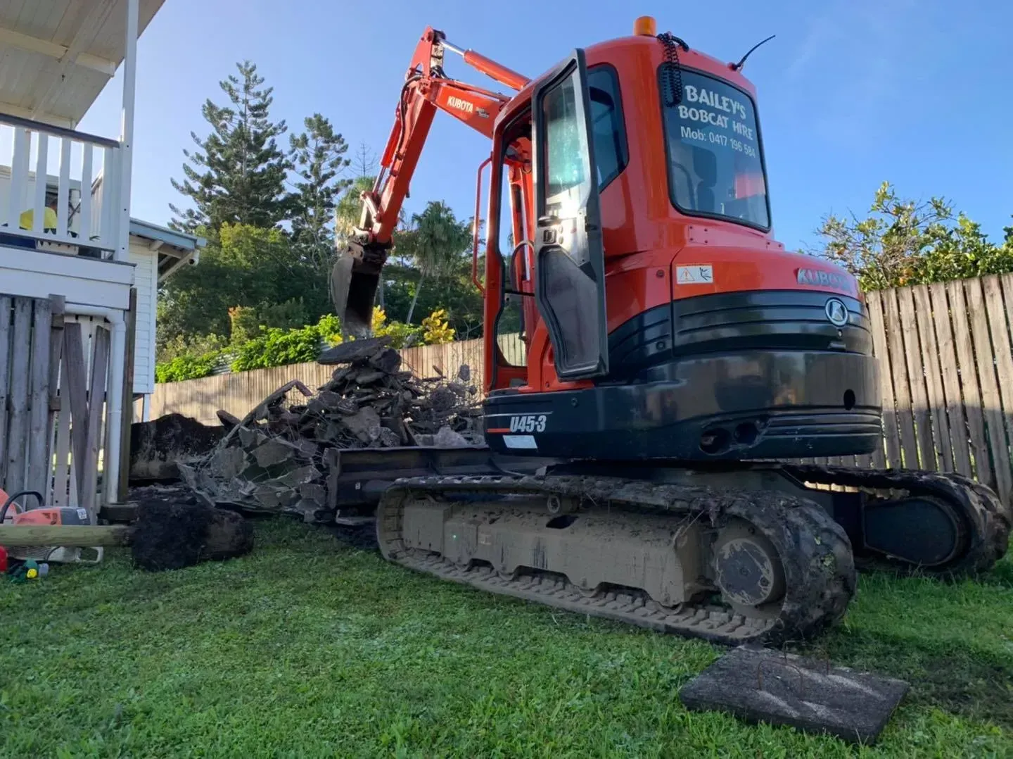 Orange Excavator Digging in A Grassy Yard Next to A House and Fence — Bailey's Bobcat & Excavator Hire In Dumbleton, QLD