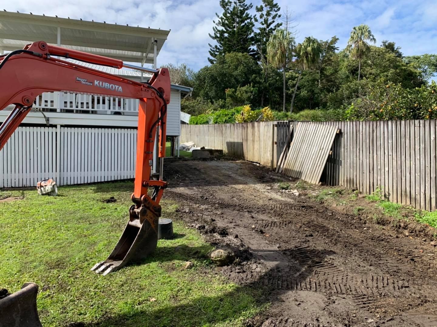 An Excavator in A Muddy Driveway Next to A White Fence and A House, Preparing the Ground — Bailey's Bobcat & Excavator Hire In Dumbleton, QLD