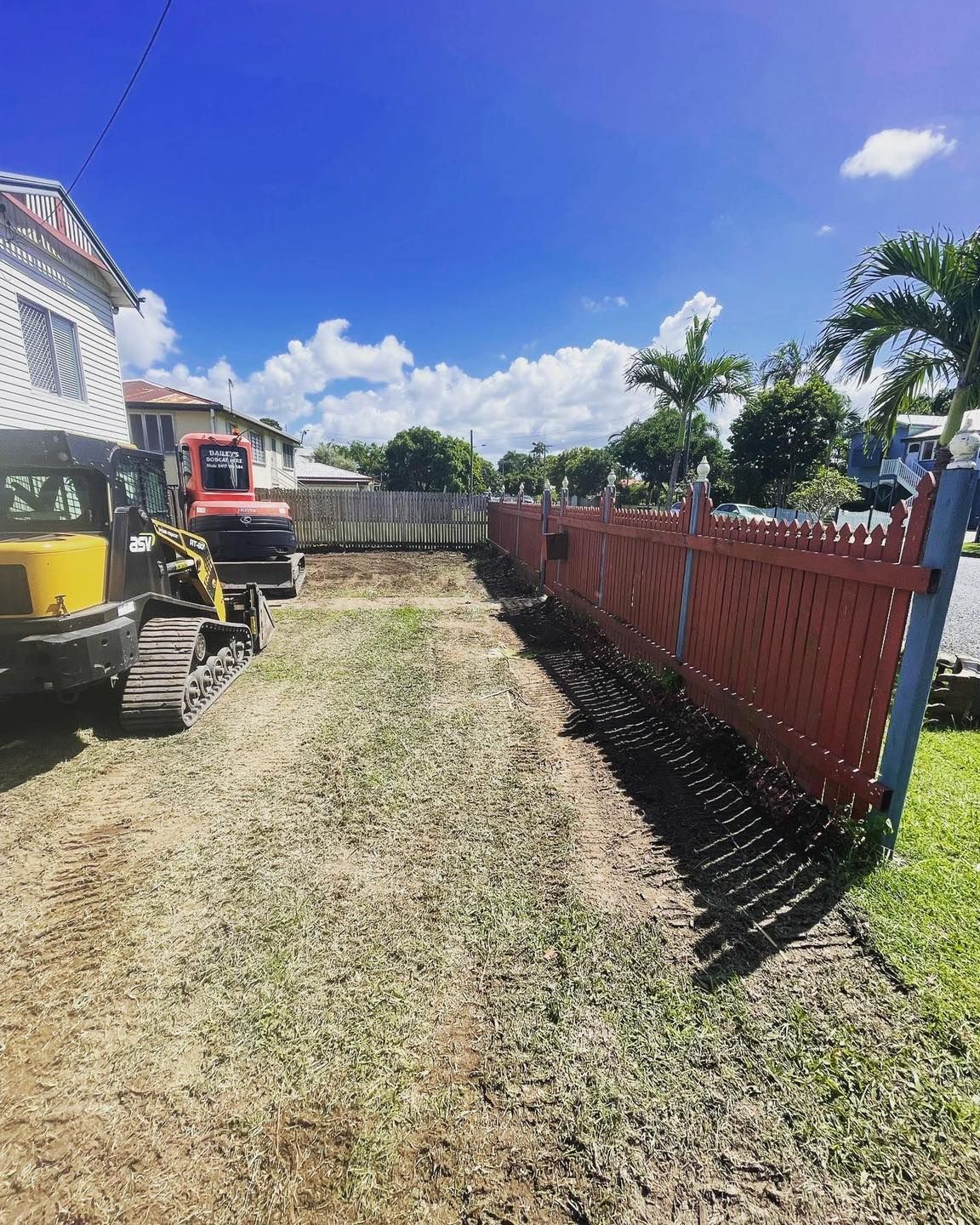 A Construction Site with A Red Fence, Dirt Patch, and Two Small Excavators Under a Blue Sky — Bailey's Bobcat & Excavator Hire In Dumbleton, QLD
