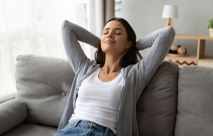 Una mujer está sentada en un sofá con los ojos cerrados y las manos detrás de la cabeza.