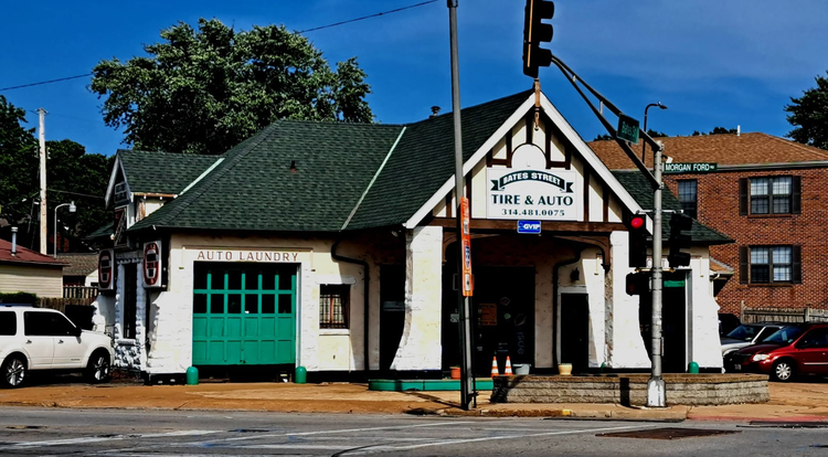 Small white building with green roof and doors at a street corner, with traffic lights and nearby cars.
