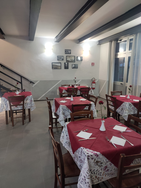 Piccola sala da pranzo di un ristorante con tovaglie rosse, sedie di legno e foto incorniciate alle pareti.