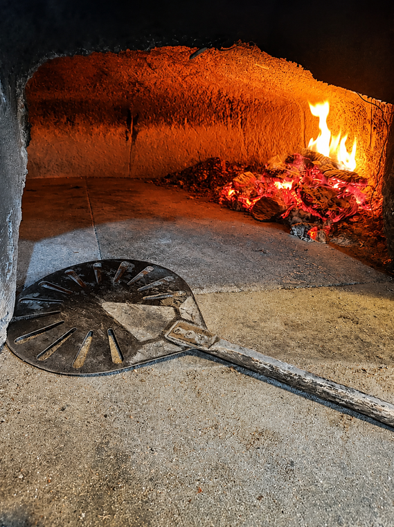 Forno a legna con fuoco acceso e pala per pizza sul pavimento in pietra.