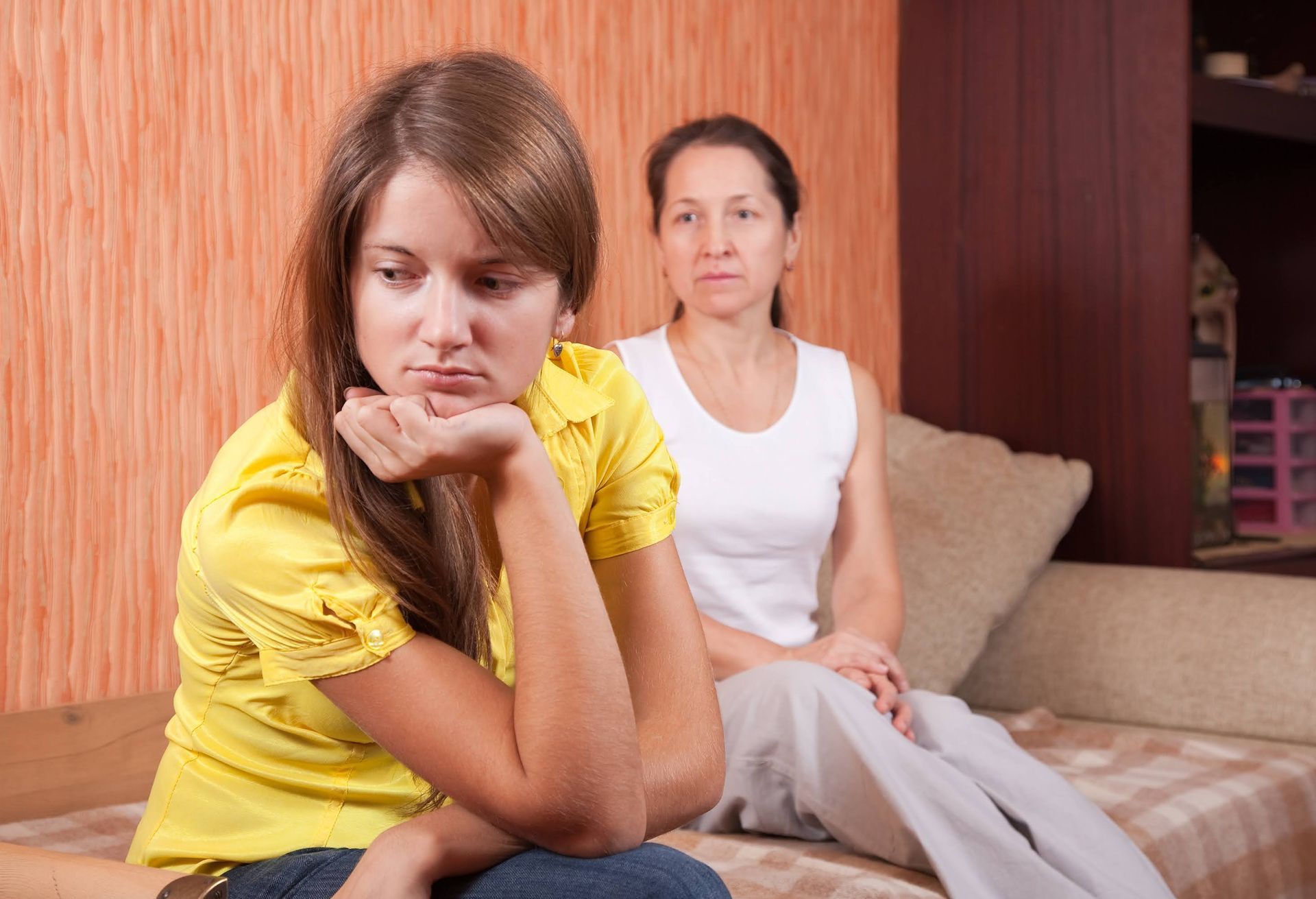 Teen girl looking sad, sits on couch with concerned mother in room with orange walls.