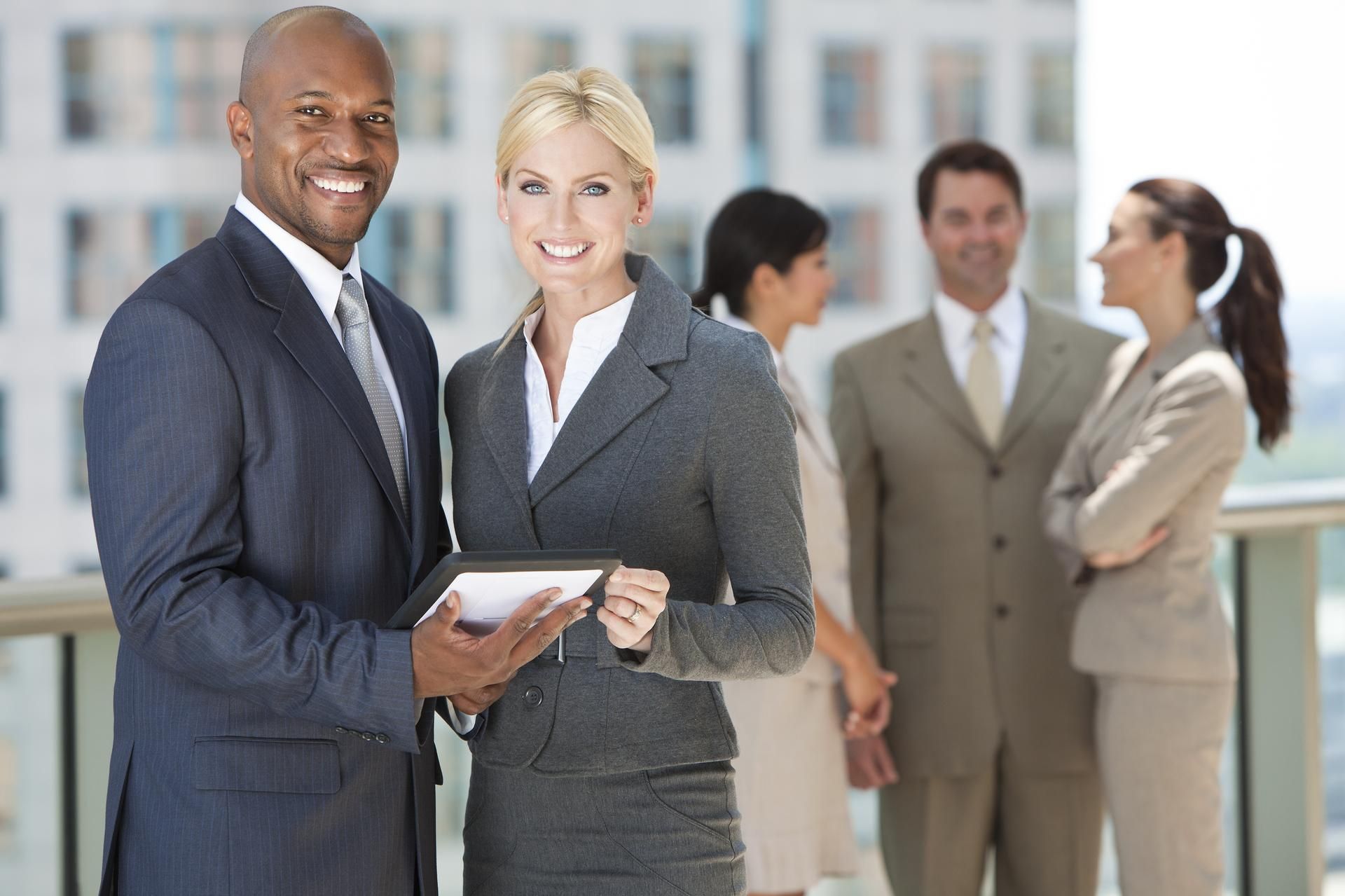 Two businesspeople smiling, holding tablet. Other colleagues blurred in the background.