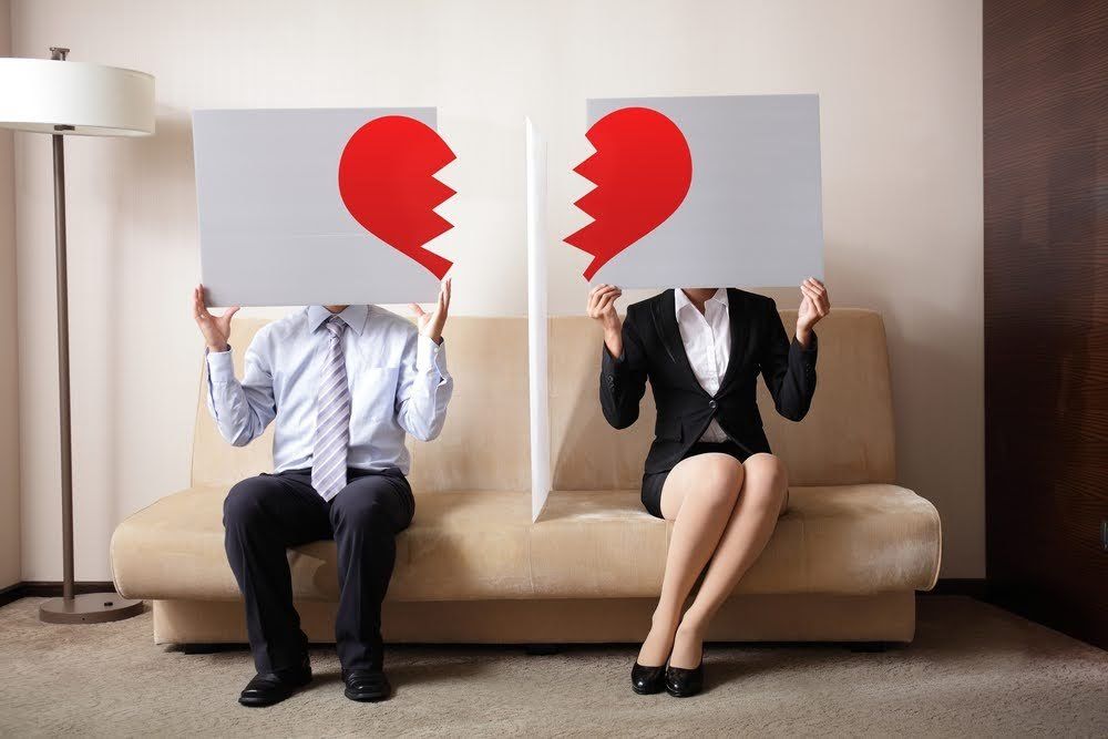 Couple holding halves of a broken heart over their faces, sitting on a couch.