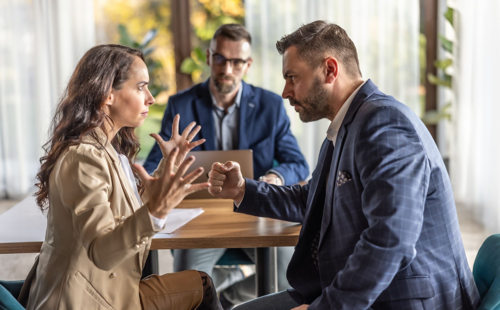 Woman and man arguing across a table; third person in background observing. Woman and man arguing across a table; third person in background observing.