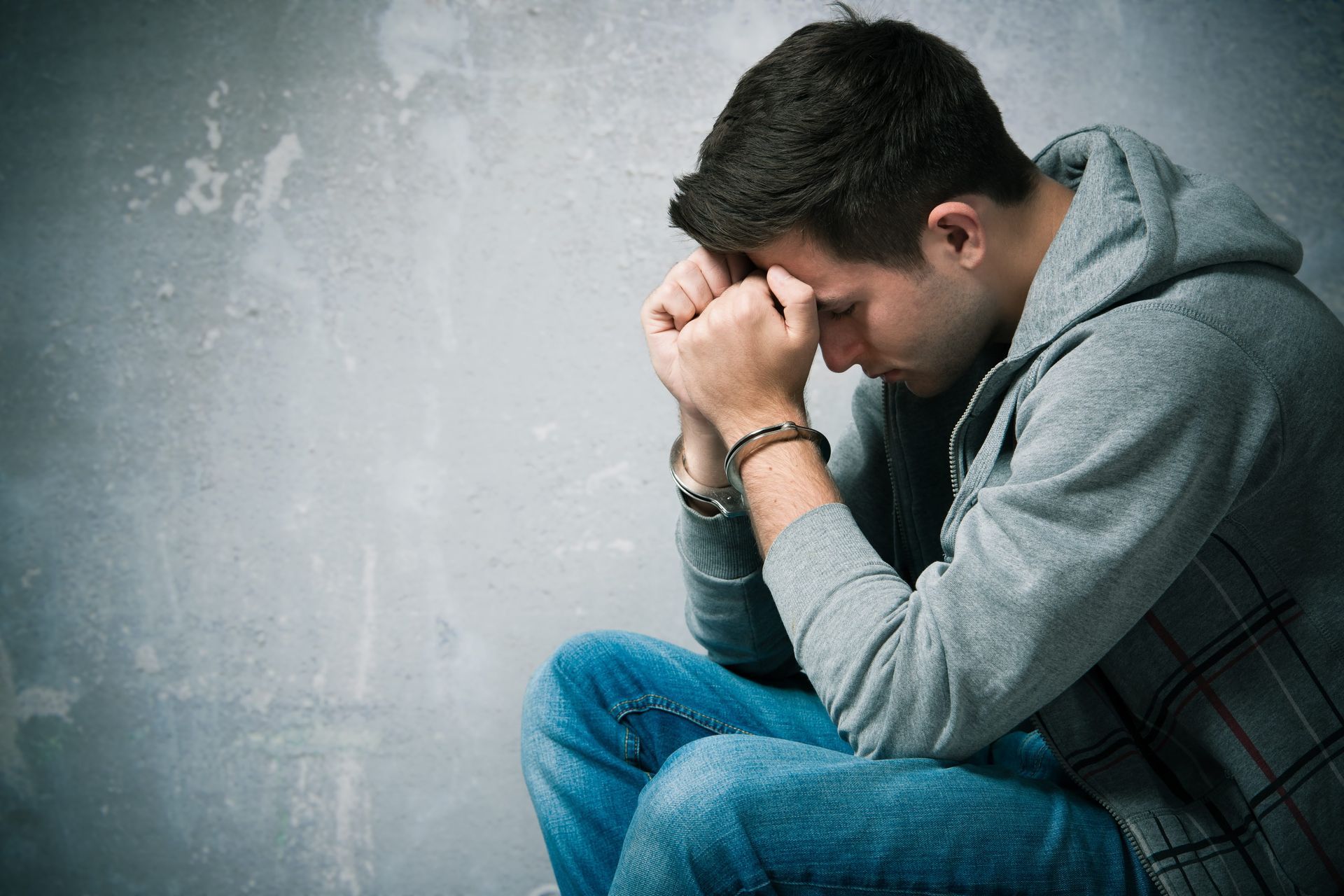 Man in a hoodie and jeans, head in hands, looking down, possibly in distress. Gray background, handcuffs visible.
