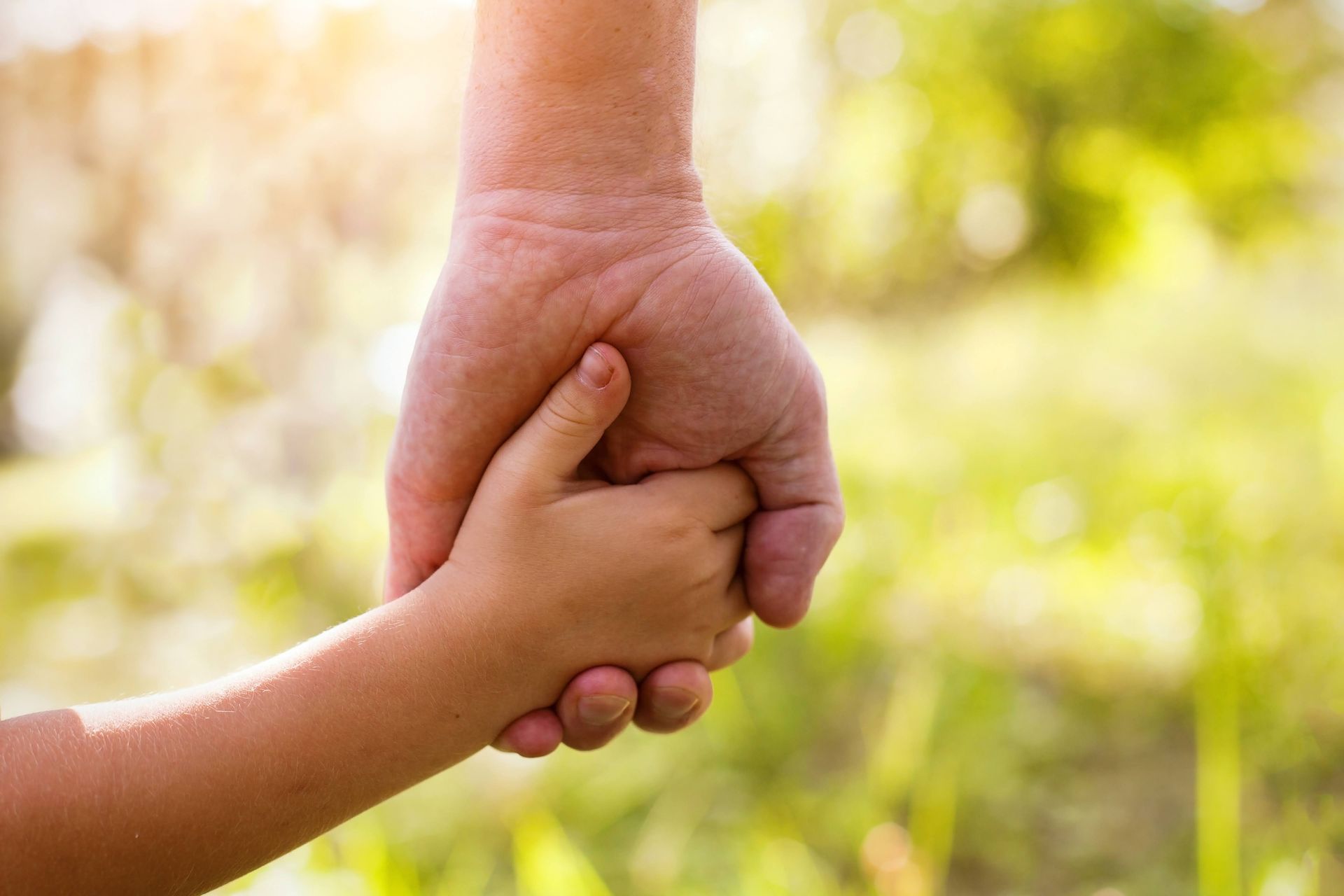 Adult hand holding a small child's hand, symbolizing care and guidance, outdoors with sunlight.