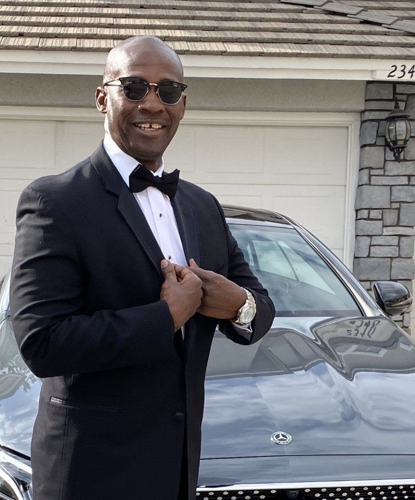 Man in a black tuxedo and sunglasses standing in front of a gray car and house.