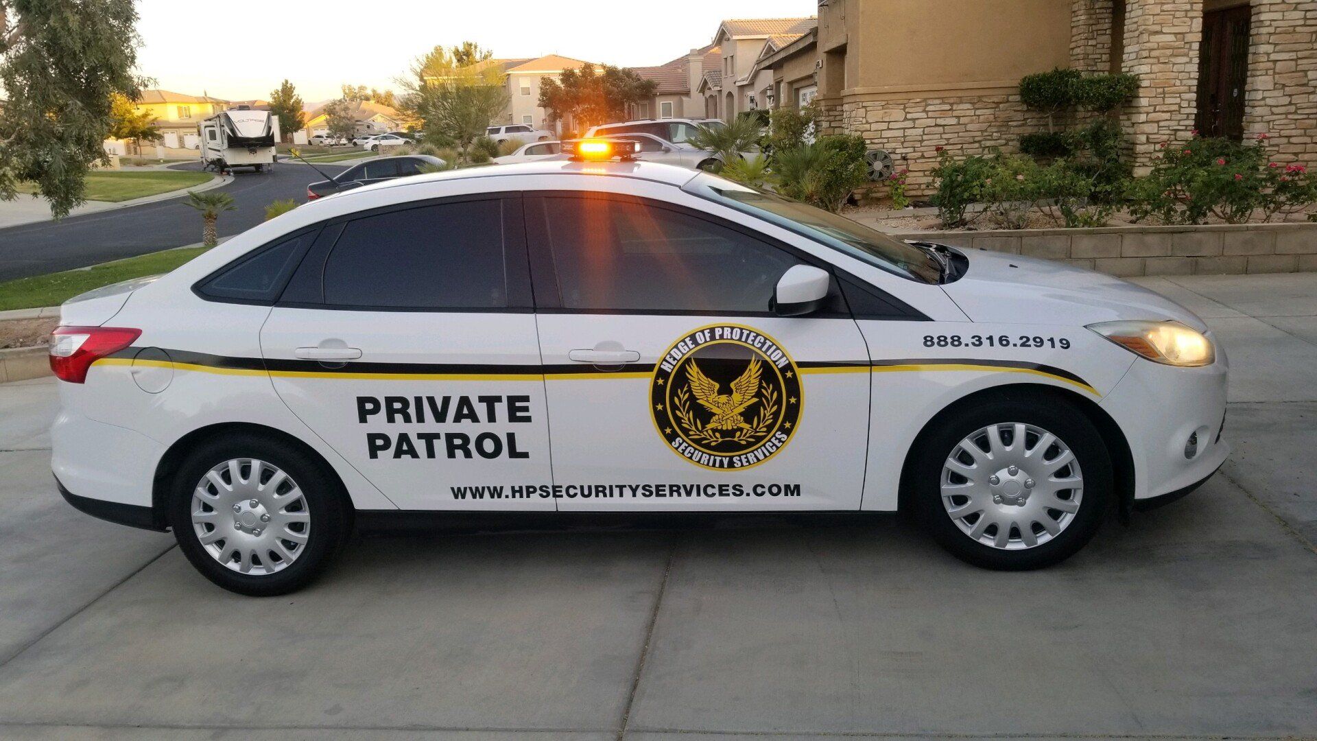 White private patrol sedan parked on a residential street at dusk, roof light flashing