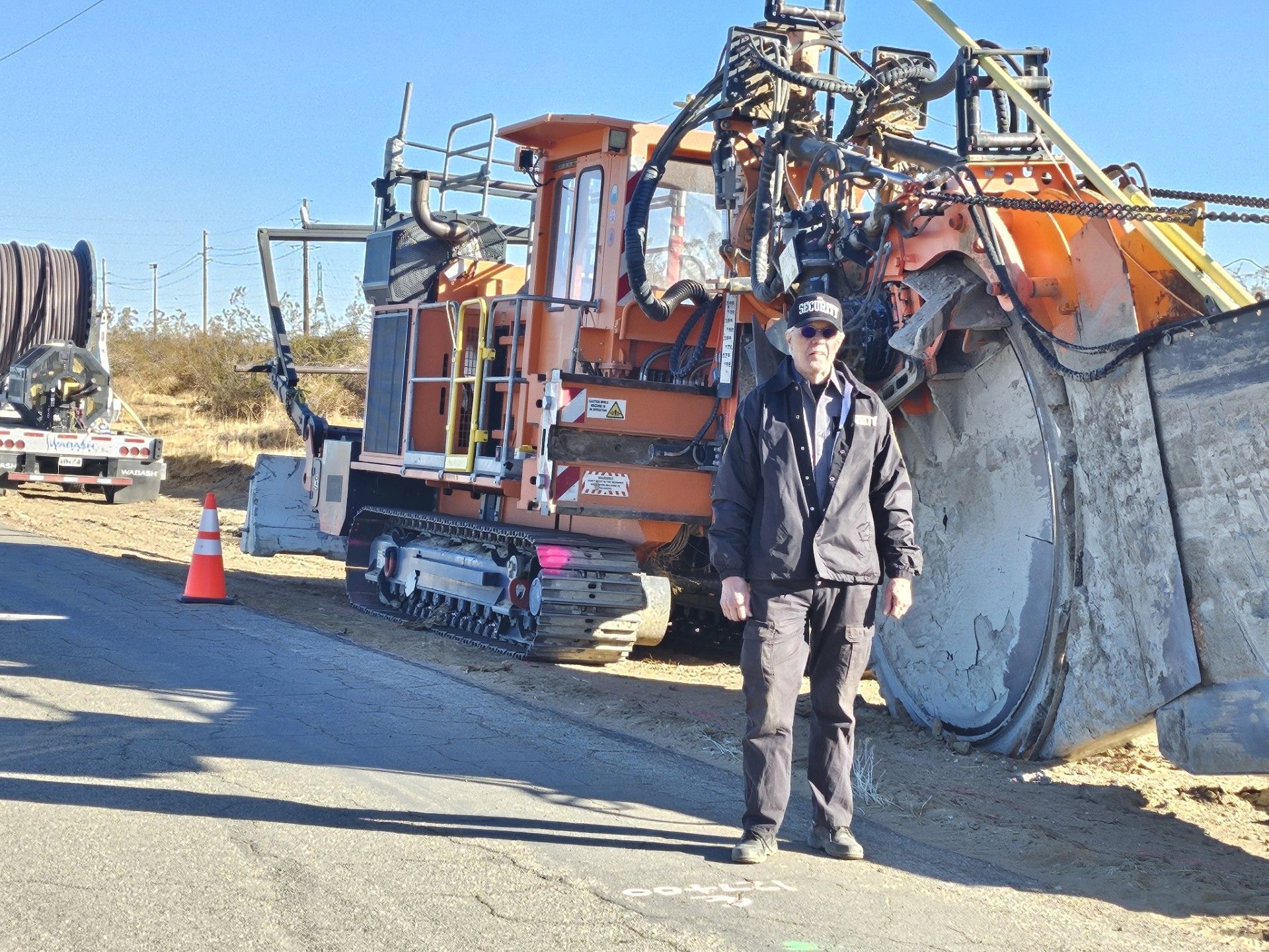 Worker standing beside an orange roadwork machine and a large concrete pipe on a roadside.