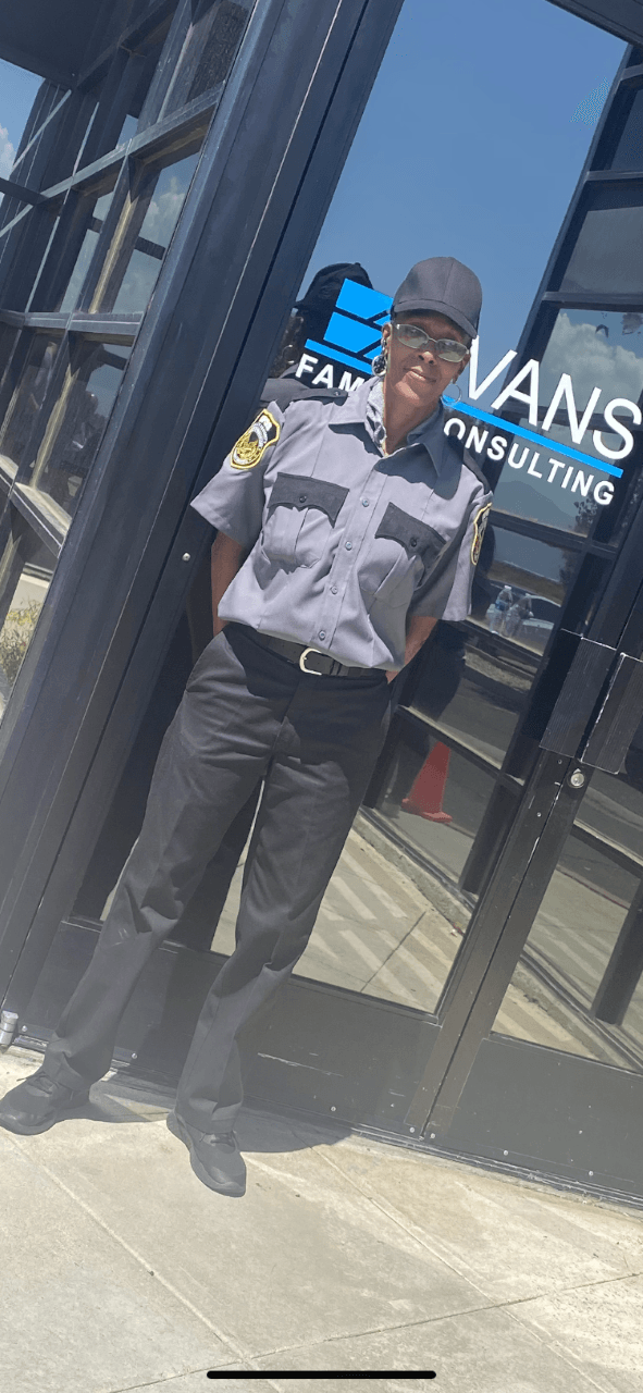 Person in gray hoodie and black cap standing outside a glass storefront in bright sunlight.