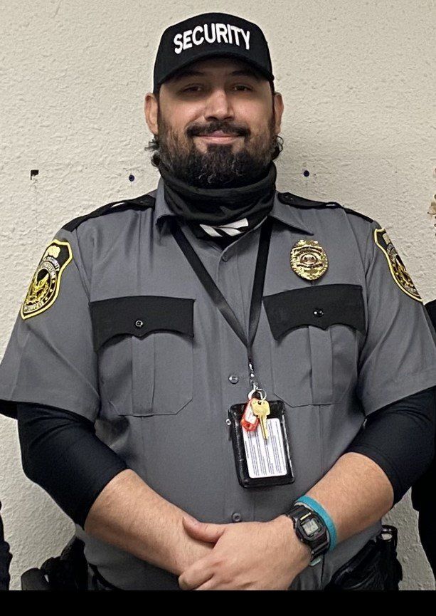 Security guard in gray uniform and black cap standing against a light wall, wearing an ID badge.