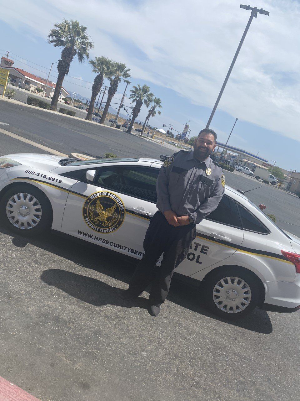 Police officer standing beside a sheriff patrol car in a sunny parking lot with palm trees and mountains in the background