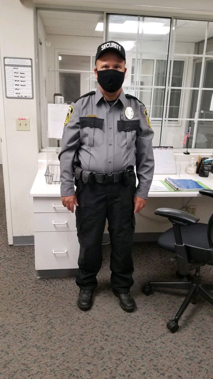 Security guard standing in an office beside a desk and chair, wearing a gray uniform and black mask.