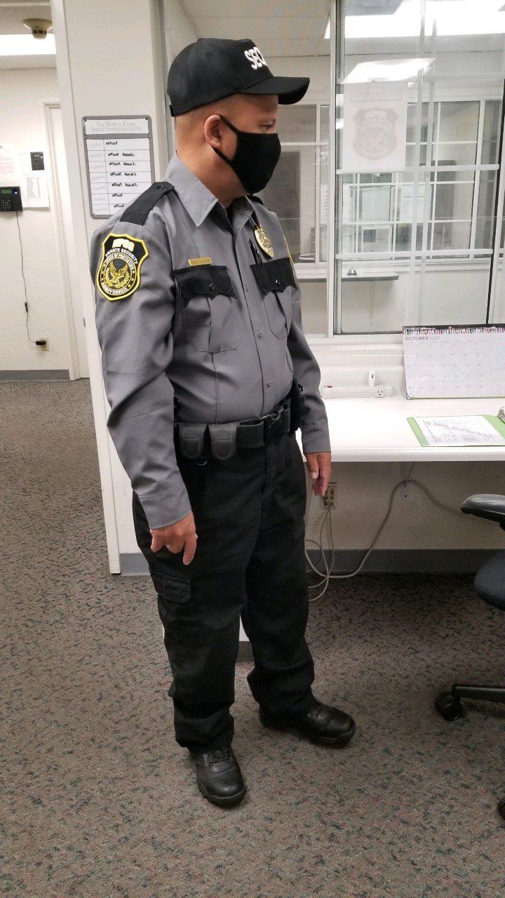 Security guard in gray uniform and black mask standing beside a table in an office hallway.