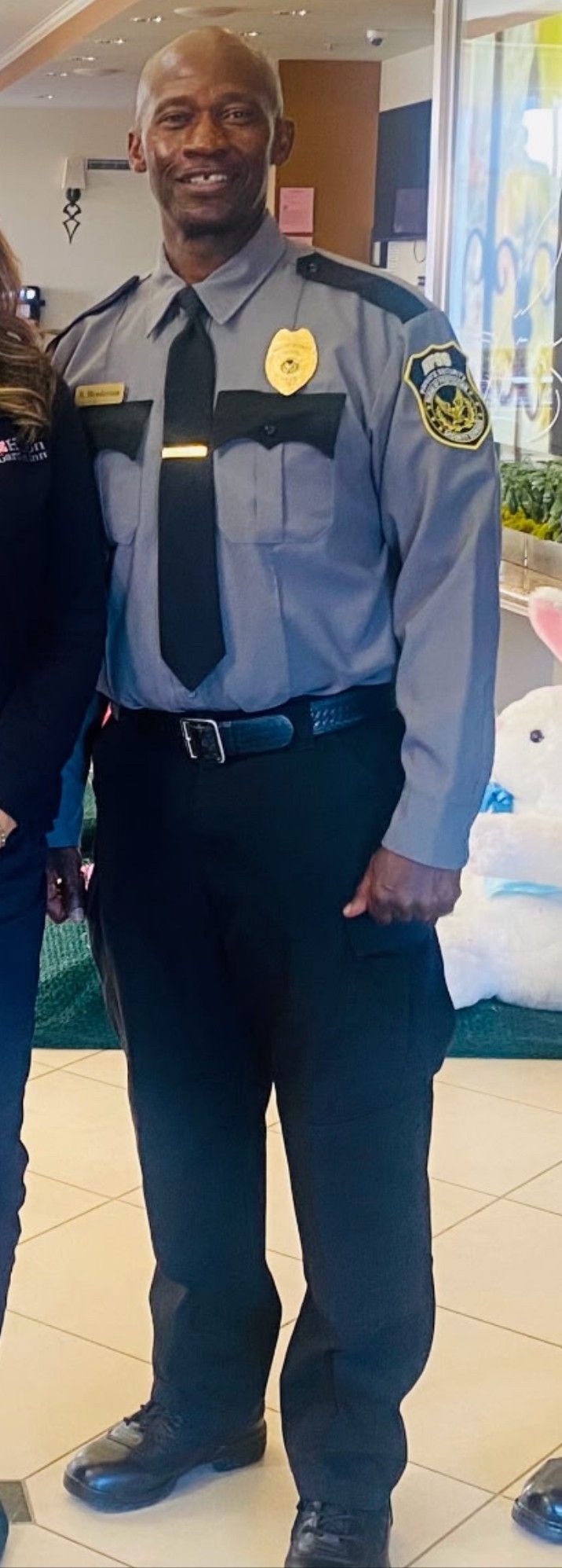 Security guard in gray uniform smiling indoors, standing with hands behind his back.