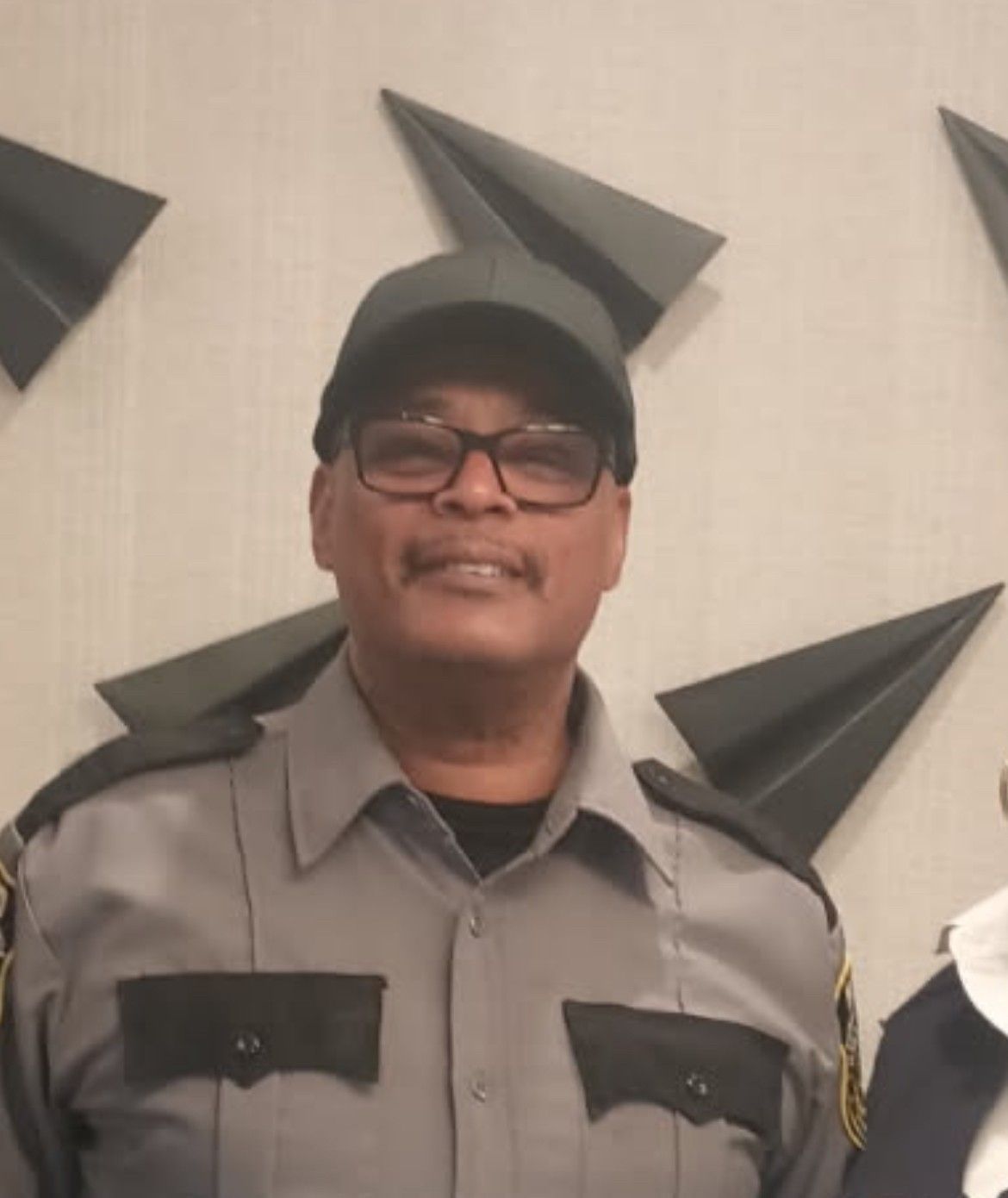 Person in a gray uniform and black cap standing indoors against a wall with black geometric decor.