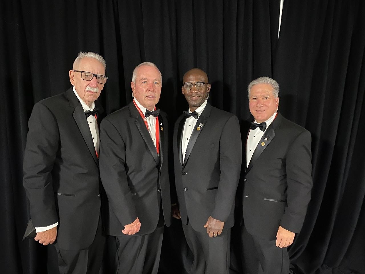Four men in black suits posing together in front of black curtains.
