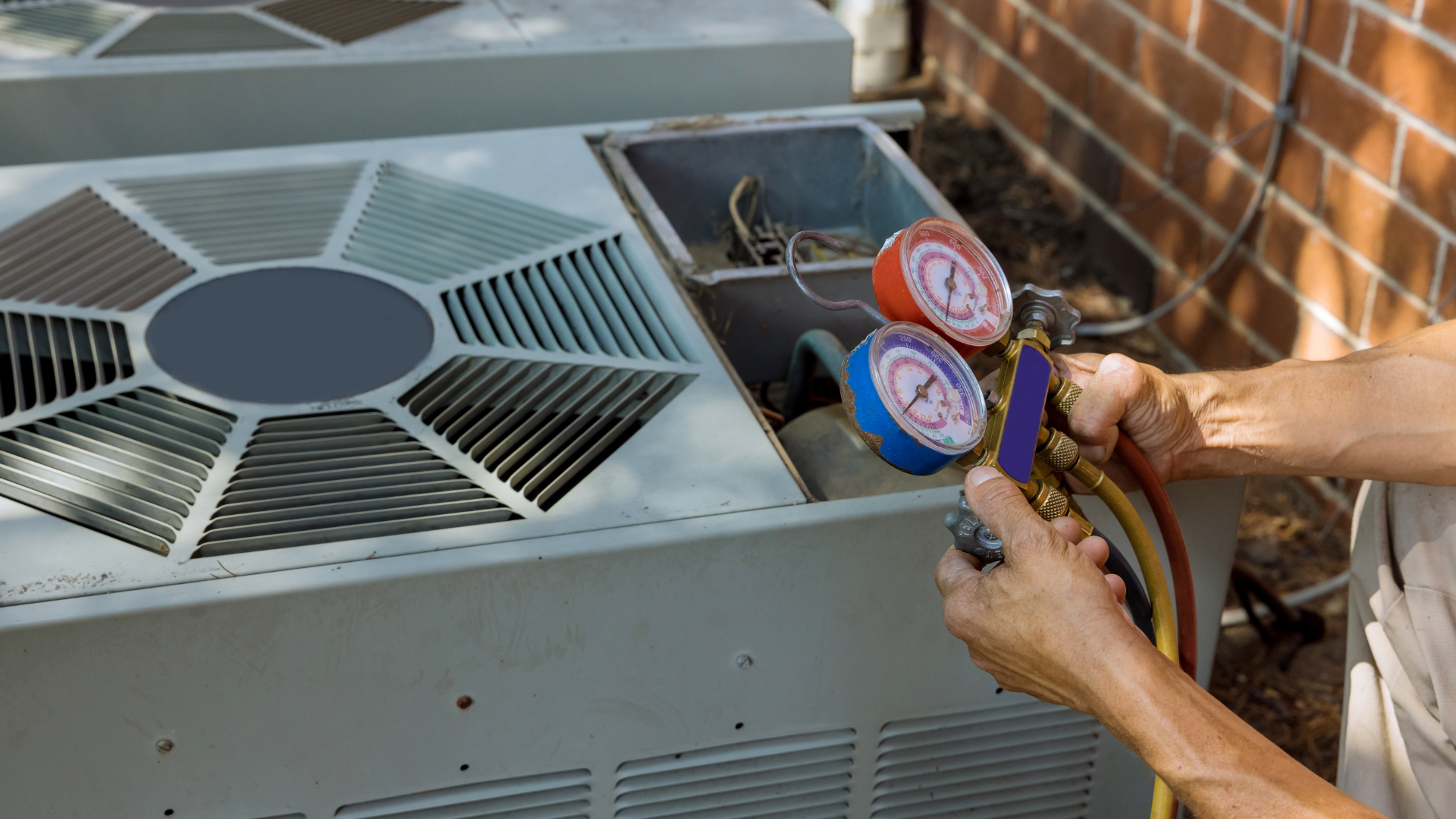 HVAC technician connecting gauges to an air conditioner. Red and blue gauges, outdoors by a brick wall.