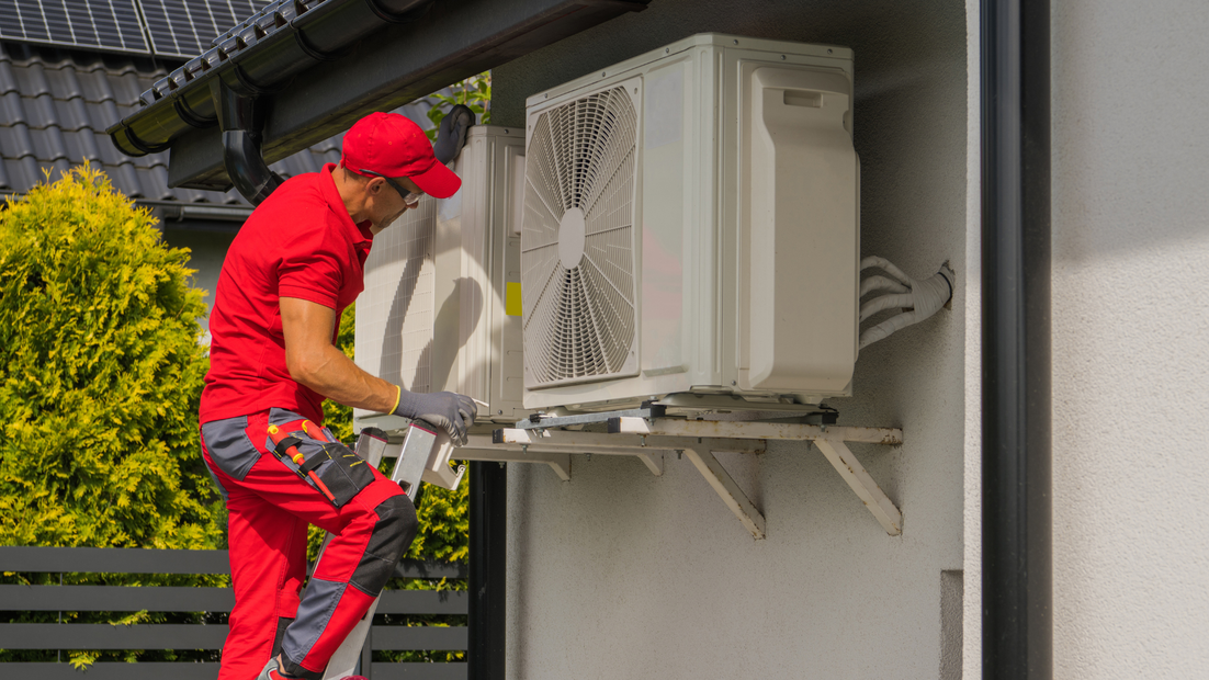 HVAC technician in red uniform repairs an air conditioning unit mounted on a wall outdoors.