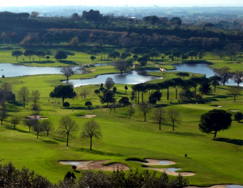 Green golf course with sand bunkers, scattered trees, and water hazards under a bright sky