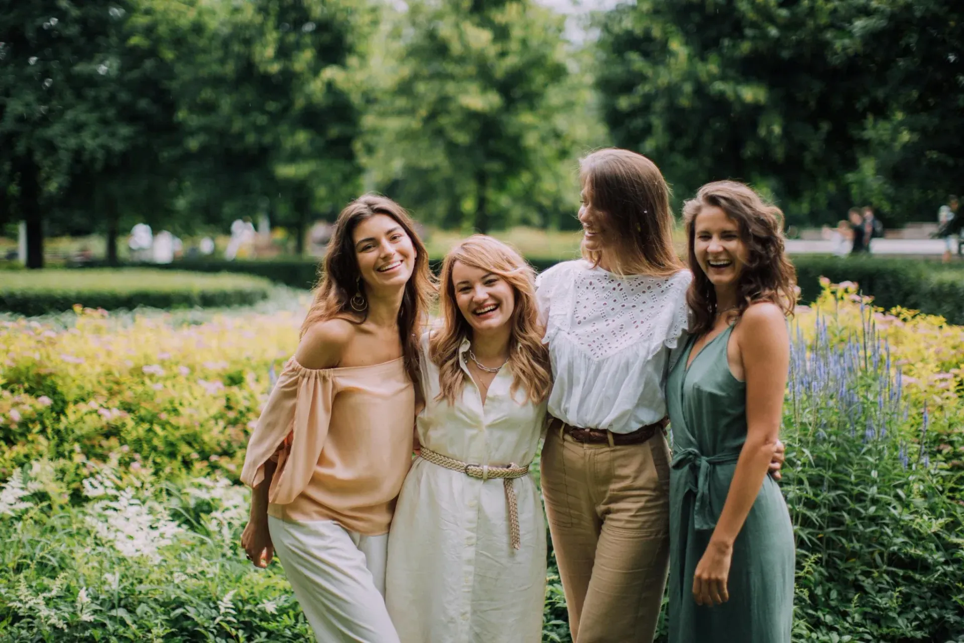 Four friends smiling in a flower-filled park, standing together in soft summer clothes.