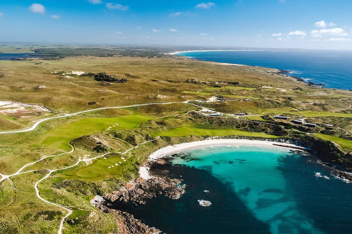 Aerial view of a turquoise cove beside green coastal cliffs and a blue ocean horizon on tasmanina golf tour