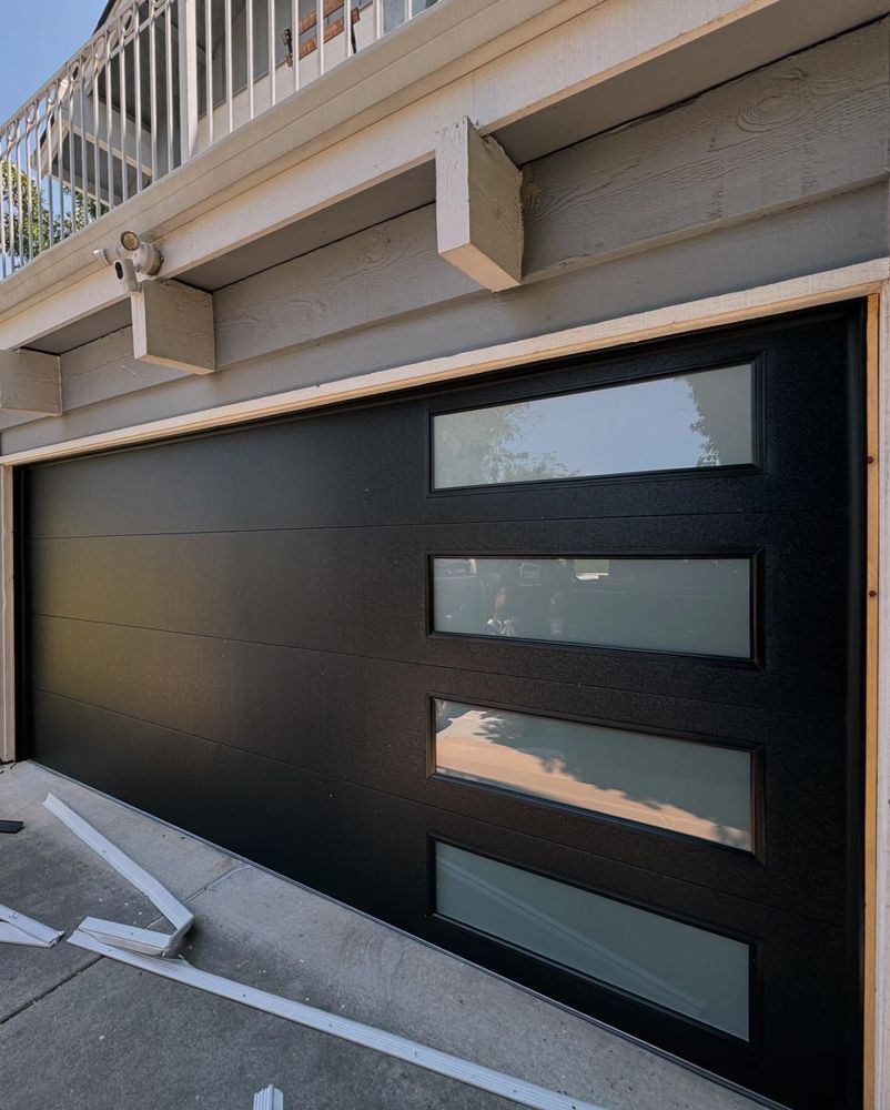 Black garage door with four horizontal windows under a balcony, beside a parking space marked by white lines Black garage door with four horizontal windows under a balcony, beside a parking space marked by white lines