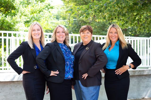 Four women in business attire posing outdoors, smiling. They stand in front of a white fence and greenery.
