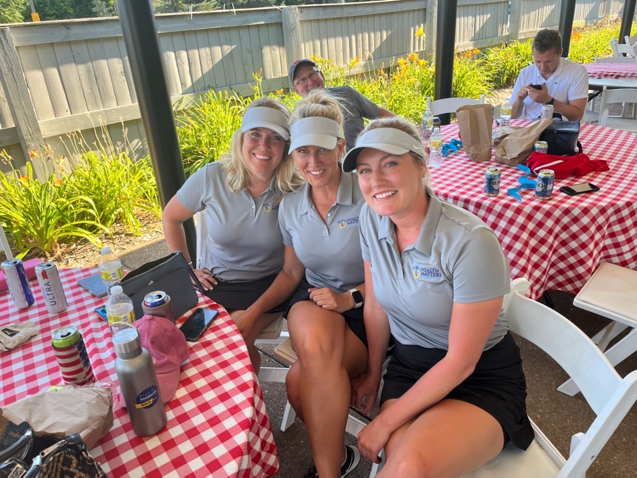 Three women in matching gray shirts and visors smile at a picnic table with a red and white checkered tablecloth.