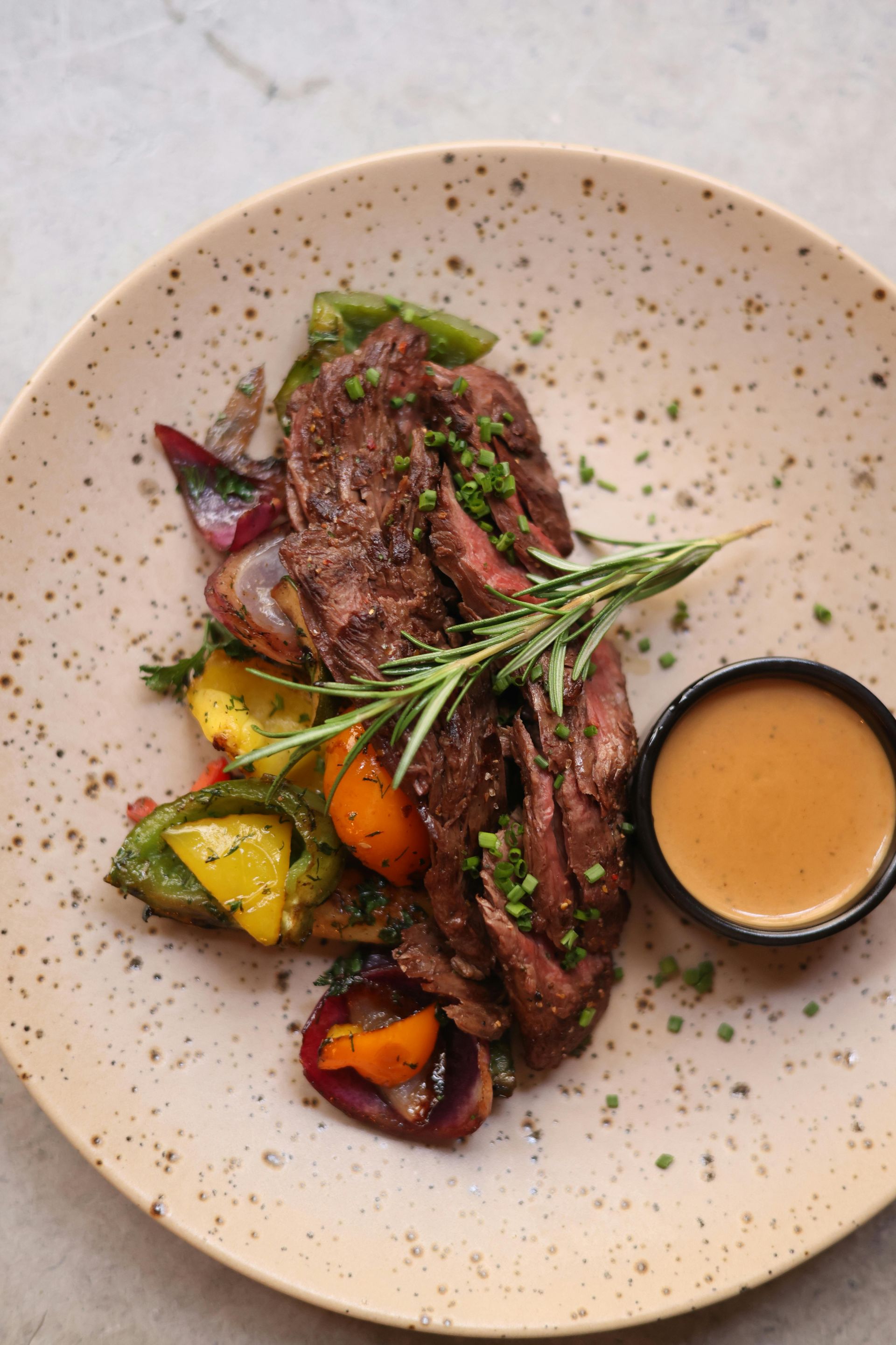 Sliced steak with grilled vegetables and a sauce in a small bowl on a speckled plate. A sprig of rosemary is on top.