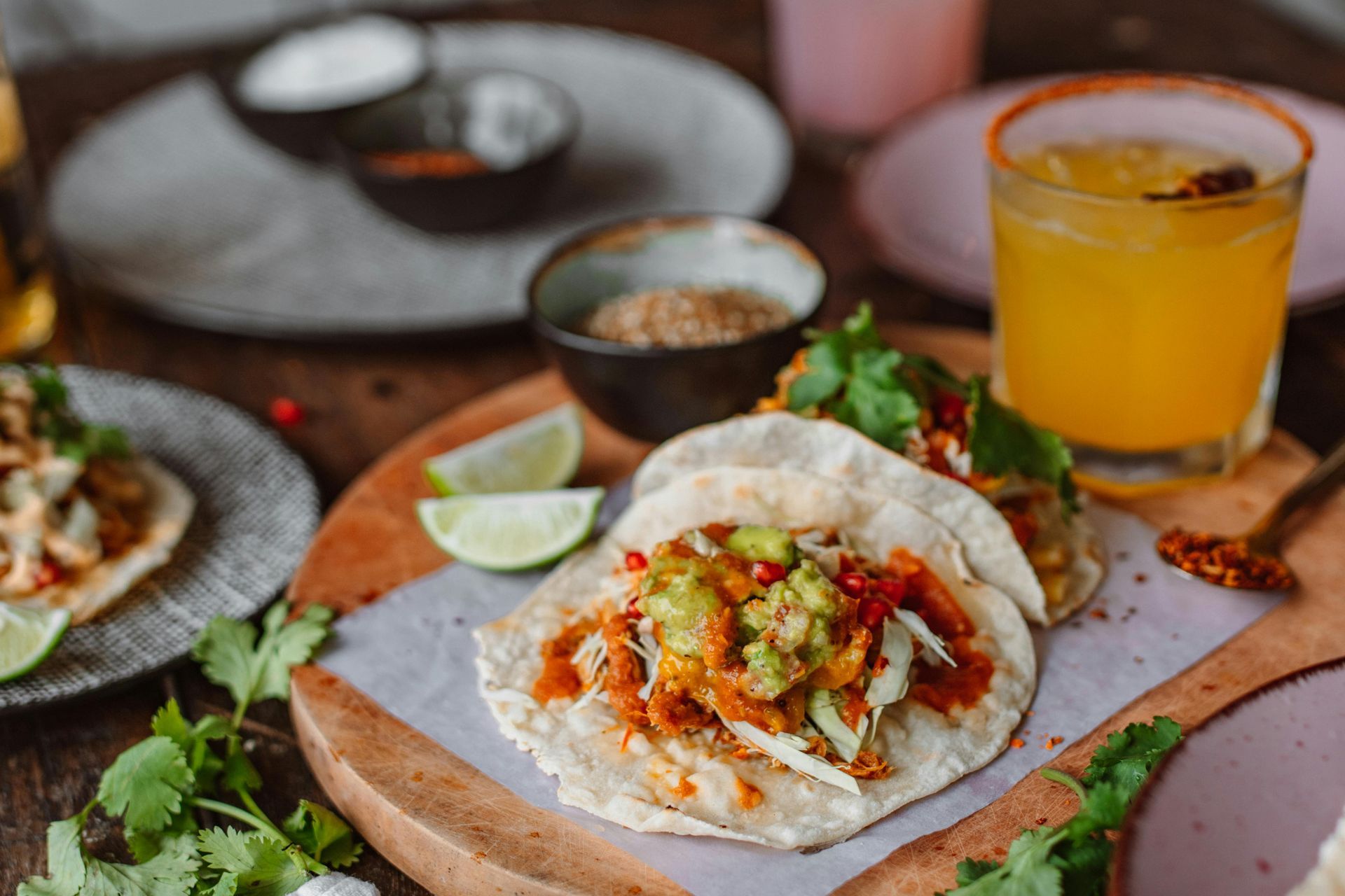 Tacos with guacamole, cilantro, and lime wedges on a wooden board, with a drink, spices, and other tacos.
