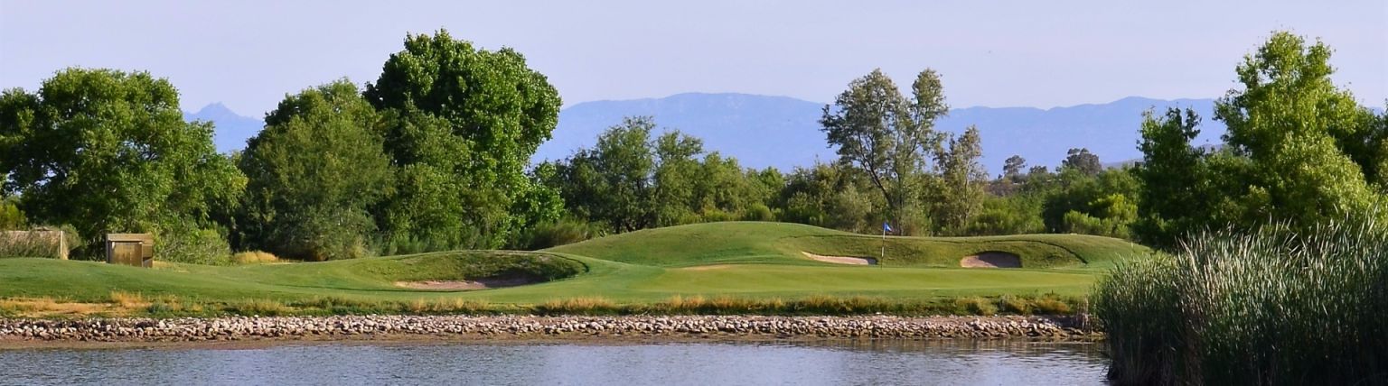 A scenic shot of a golf course along a body of water. Lush green trees and grass line the landscape under a blue sky.