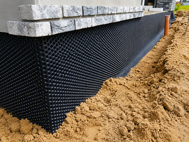 Gravel-filled trench with a white pipe running beside a building foundation on a dirt slope