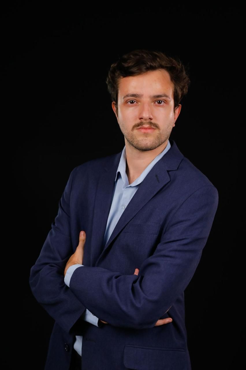 Um homem de terno está de pé com os braços cruzados em frente a um fundo preto.