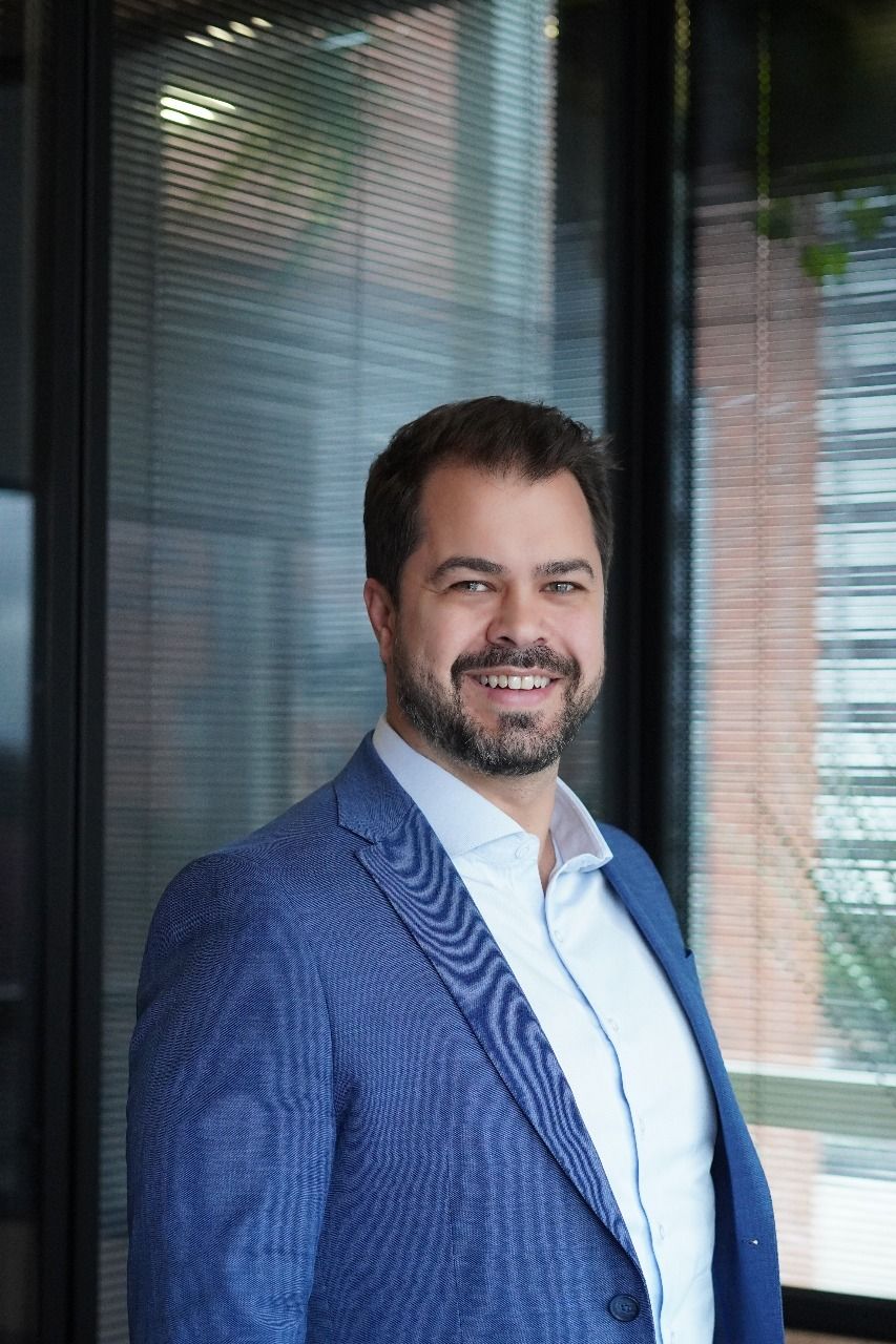 Um homem de terno azul e camisa branca está parado em frente a uma janela.
