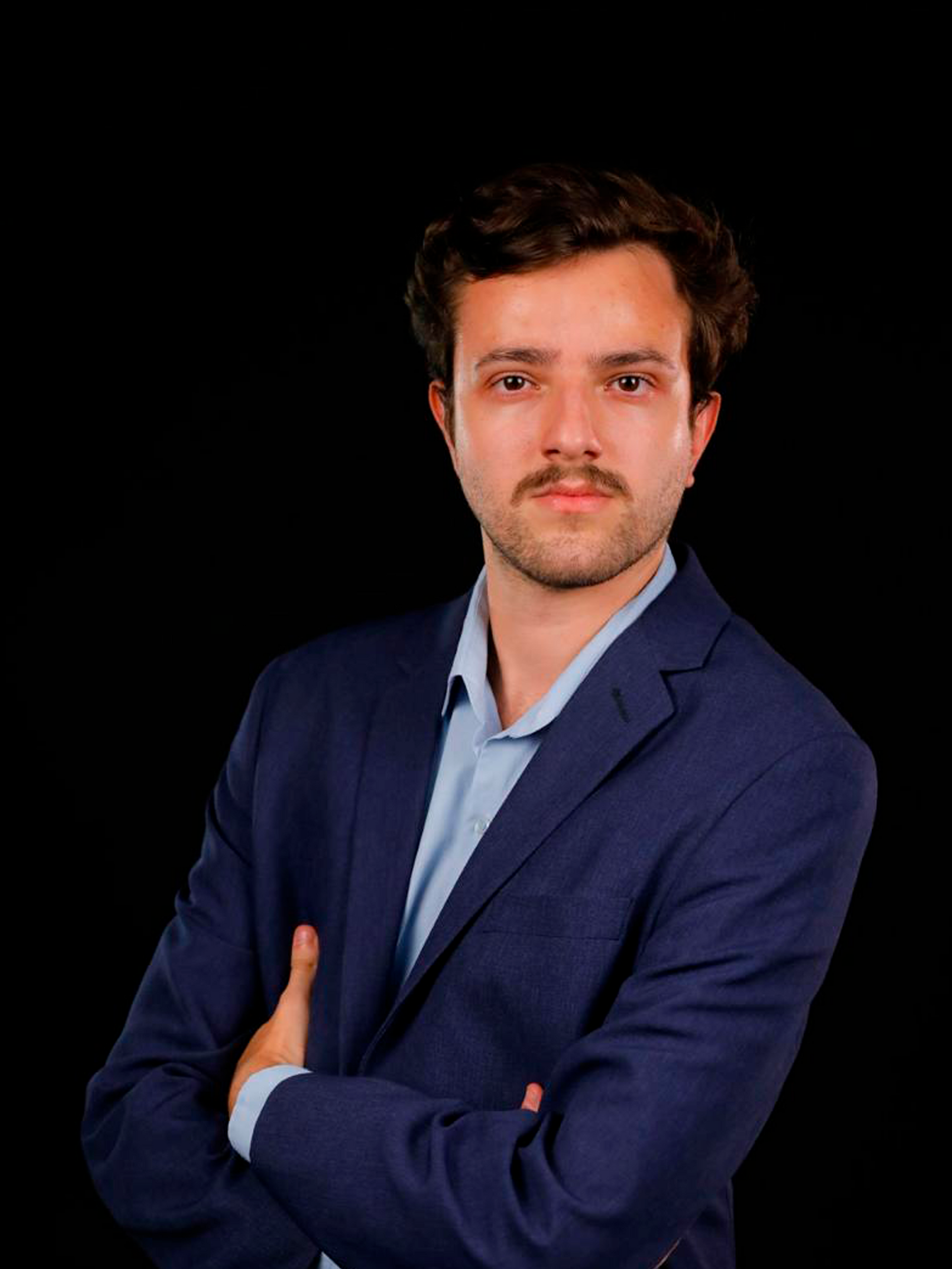 Um homem de terno está de pé com os braços cruzados em frente a um fundo preto.