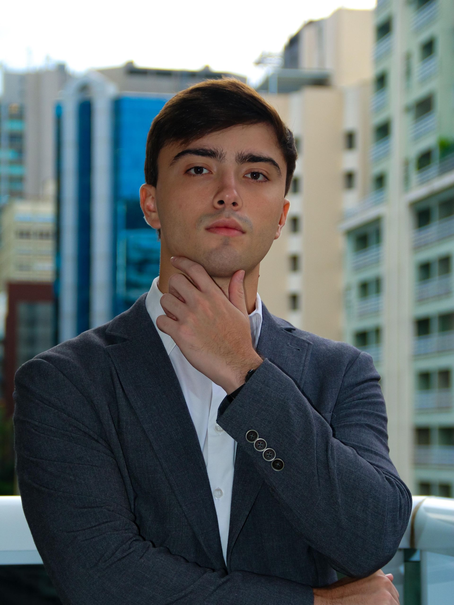 Um homem de terno está parado em frente a um prédio com a mão no queixo.