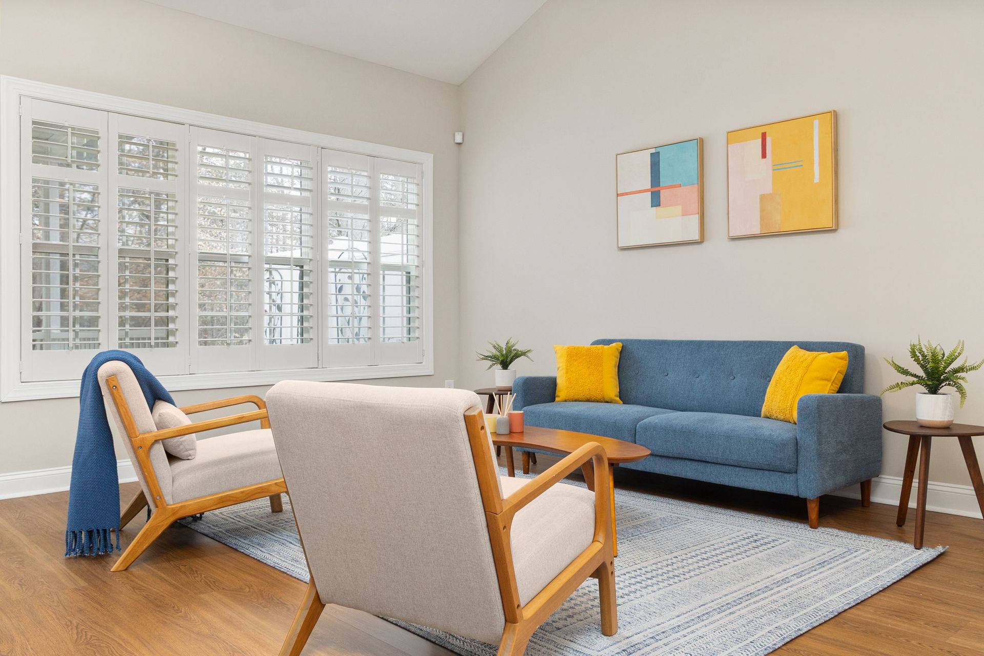 Bright living room with blue sofa, wooden chairs, rug, plants, and wall art by a window