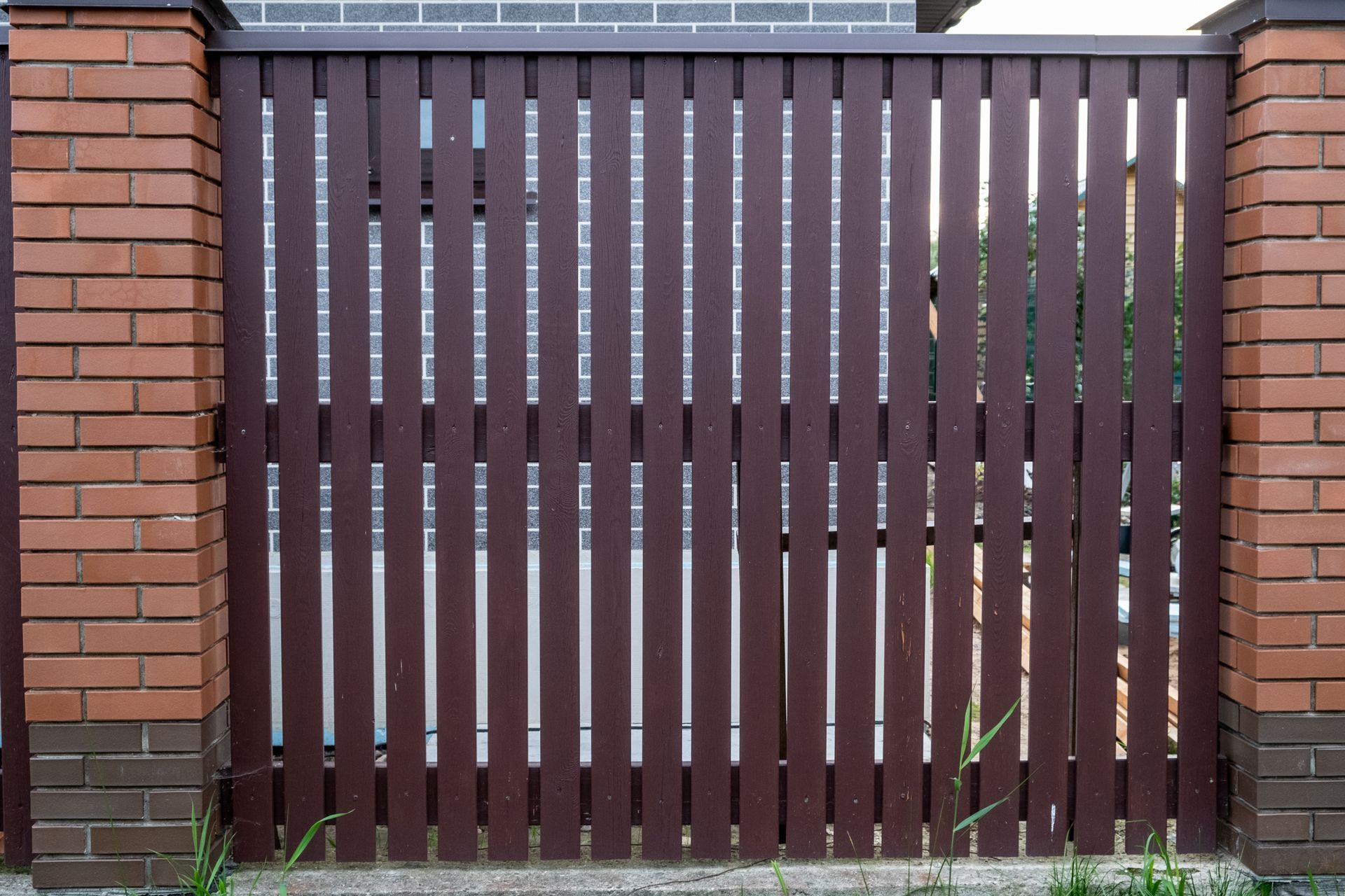 Brown wooden gate between brick pillars and a metal fence with slats behind it