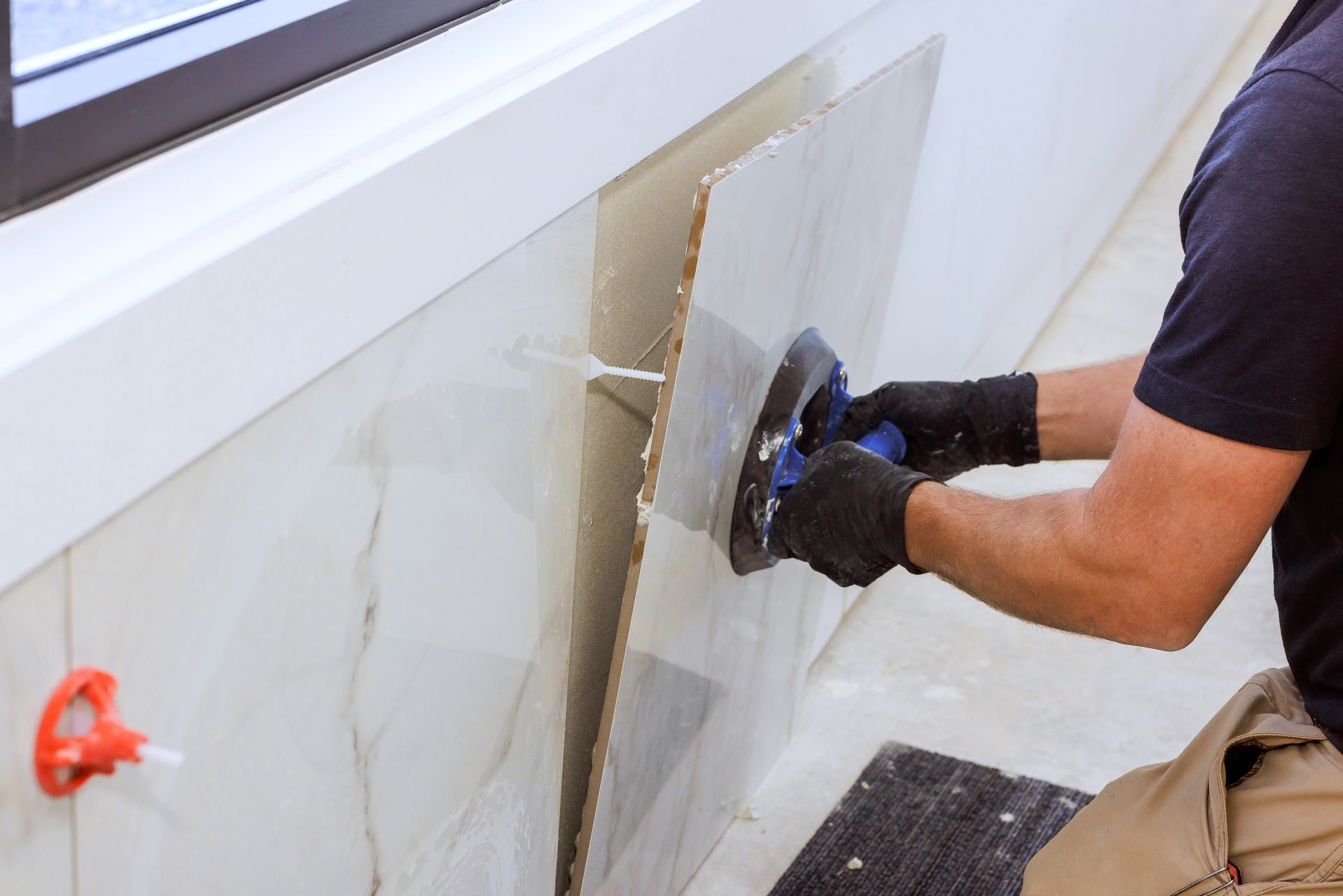 Worker smoothing wet plaster on a white wall with a trowel, wearing black gloves and kneeling on a floor mat