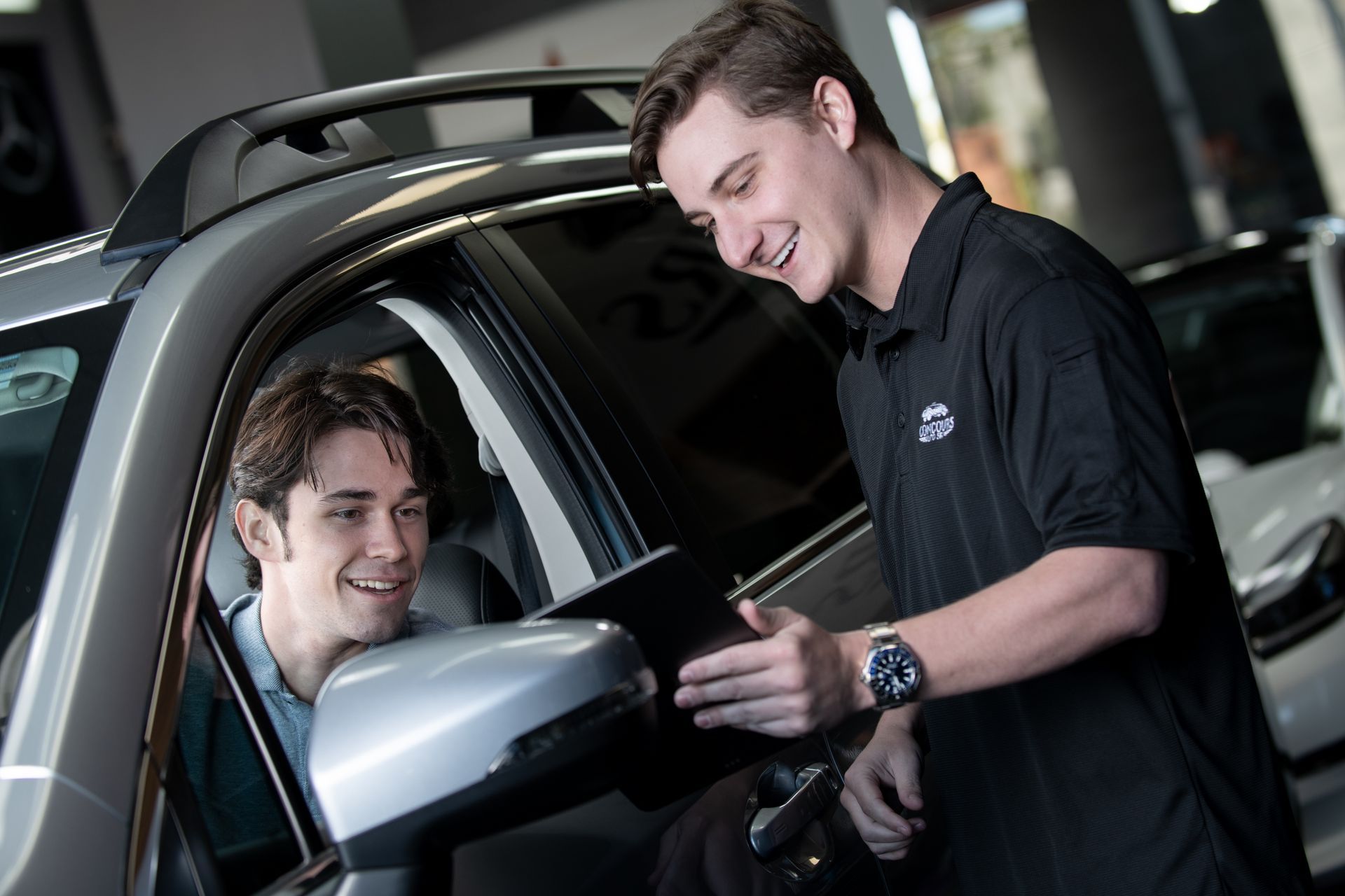 Man in car smiles at another man, both in a car dealership, discussing the side mirror.